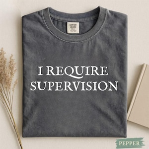 May include: Dark grey Comfort Colors t-shirt with the white text "I REQUIRE SUPERVISION". The folded t-shirt has a crew neck and a small tag near the neckline. The background is neutral. The t-shirt is a casual wear item.