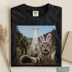 May include: Black t-shirt with a colorful graphic featuring a cat wearing bunny ears taking a selfie with a Bigfoot, two rabbits, and a UFO. The image includes a rainbow beam of light and a basket of Easter eggs.
