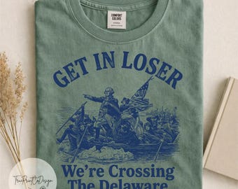 Può includere: Una t-shirt verde salvia con una stampa grafica blu scuro di George Washington che attraversa il fiume Delaware. Il testo sopra l'illustrazione recita GET IN LOSER e il testo sotto We're Crossing The Delaware. La maglietta ha un girocollo.