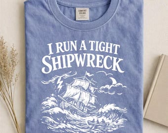 Può includere: T-shirt blu jeans con la scritta bianca "I RUN A TIGHT SHIPWRECK". La grafica raffigura una nave in una tempesta con onde che si infrangono. La maglietta ha un girocollo e un'etichetta Comfort Colors.