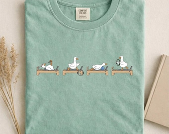 Può includere: T-shirt verde chiaro con una grafica di anatre dei cartoni animati che si esercitano su attrezzature per il Pilates. La maglietta ha uno scollo tondo e un'etichetta con la scritta "Comfort Colors". La parola "BAY" è stampata in basso a destra.