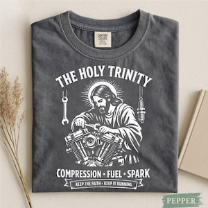 Può includere: T-shirt grigio scuro con grafica bianca. La grafica presenta un'illustrazione di Gesù che lavora su un motore, con la scritta "THE HOLY TRINITY" sopra e "COMPRESSION FUEL SPARK" sotto. La maglietta ha anche la scritta "KEEP THE FAITH - KEEP IT RUNNING".