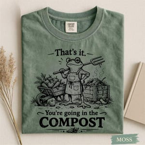 May include: Moss green t-shirt featuring a frog character wearing glasses and an apron, holding a garden fork. The shirt has the text "That's it. You're going in the COMPOST" in black. A wheelbarrow and compost bin are also in the graphic.