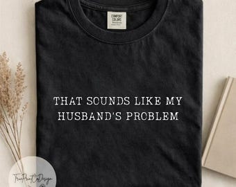 Può includere: Una t-shirt nera a girocollo con il testo bianco "THAT SOUNDS LIKE MY HUSBAND'S PROBLEM" stampato in un carattere in stile macchina da scrivere al centro del petto. La maglietta presenta un'etichetta del marchio visibile nel colletto.