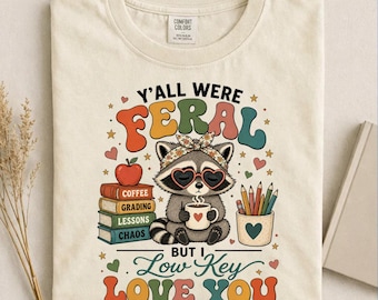 Può includere: T-shirt color avorio con grafica di procione con occhiali a forma di cuore e fascia. Il testo recita "Y'all Were Feral But I Low Key Love You". Il design include libri, una mela, matite e stelle.