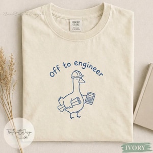 Może przedstawiać: Kremowy t-shirt z krótkim rękawem z niebieską grafiką liniową przedstawiającą gęś w kasku ochronnym, trzymającą zwinięty projekt i kalkulator. Tekst nad gęsią brzmi "off to engineer" i jest napisany prostą, niebieską czcionką.