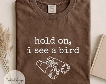 Può includere: Una t-shirt marrone a maniche corte con scollo rotondo. Sul davanti è presente la scritta bianca "hold on, i see a bird" sopra un'illustrazione a linee bianche di un binocolo. L'etichetta interna sul collo riporta "Comfort Colors".