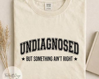 Può includere: Una t-shirt a maniche corte color avorio con testo nero che recita UNDIAGNOSED in un grande carattere universitario arcuato, seguito dal testo BUT SOMETHING AIN'T RIGHT con una stella nera su ciascun lato, stampata su una t-shirt in cotone.