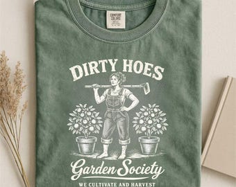 Può includere: T-shirt verde muschio con la scritta "DIRTY HOES" sopra una grafica di una donna con una zappa, con le parole "Garden Society" e "WE CULTIVATE AND HARVEST" sotto. Due piante in vaso affiancano la donna.