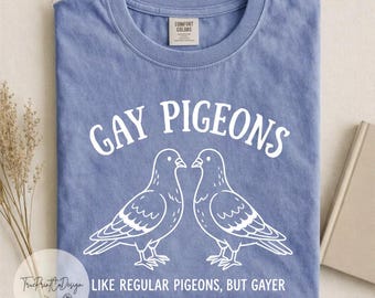 Può includere: Una t-shirt blu a girocollo con una grafica bianca di due piccioni l'uno di fronte all'altro. Il testo sopra i piccioni recita GAY PIGEONS e il testo sottostante recita LIKE REGULAR PIGEONS, BUT GAYER. La maglietta ha un colletto a coste e maniche corte.