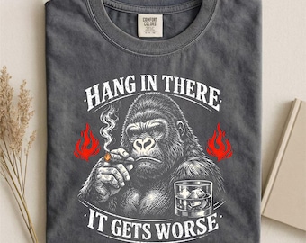 Può includere: T-shirt grigio scuro con un gorilla che fuma un sigaro e tiene in mano un bicchiere di whisky. Il testo recita "Hang in There, It Gets Worse" in bianco con dettagli di fiamme rosse.