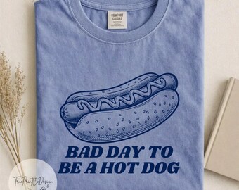 Può includere: Una t-shirt azzurra a girocollo con un'illustrazione a linee blu scuro di un hot dog con senape. Il testo "BAD DAY TO BE A HOT DOG" è stampato in lettere maiuscole blu scuro sotto la grafica. La maglietta presenta un'etichetta sul collo.