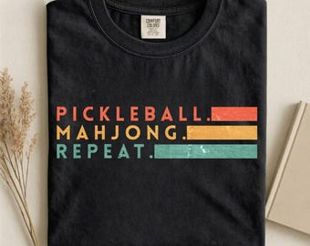 Può includere: T-shirt nero con le parole "PICKLEBALL. MAHJONG. REPEAT." in arancione, giallo e turchese. La maglietta ha un girocollo e un'etichetta con la scritta "COMFORT COLORS."