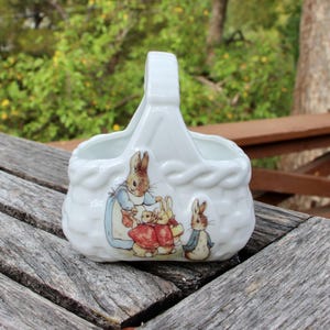May include: A white ceramic basket with a woven texture and a central handle. The front features a colorful illustration of Peter Rabbit characters, including a rabbit in a blue coat and others in red clothing, depicted in a classic storybook style.