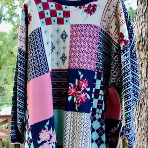May include: A long-sleeve knit sweater featuring a multi-colored patchwork design. The pattern includes various geometric shapes, floral motifs, and checkered squares in shades of navy blue, pink, light blue, and cream, finished with a ribbed crew neckline.