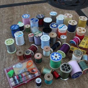 May include: A collection of sewing supplies, including spools of thread in various colors, a small sewing kit with scissors, and a measuring tape. The spools are in shades of blue, green, red, and yellow. A variety of sizes are visible.
