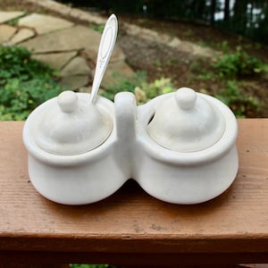 May include: A white ceramic salt and pepper set with a handle, two compartments, and matching lids. A silver spoon rests in one compartment. The set is on a wooden surface, with a blurred green background.