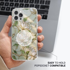 May include: Clear phone case with a floral design featuring white magnolia flowers and green leaves. The case is easy to hold and compatible with Popsockets. The design has gold accents and is set against a light green background.