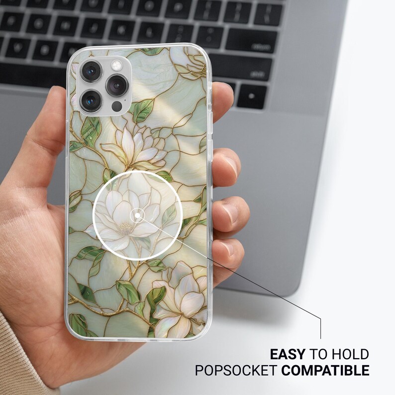 May include: Clear phone case with a floral design featuring white magnolia flowers and green leaves. The case is easy to hold and compatible with Popsockets. The design has gold accents and is set against a light green background.