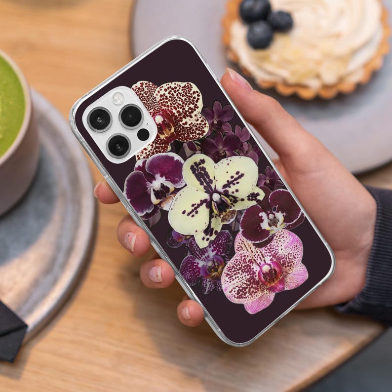 May include: A clear phone case with a floral design of orchids in shades of purple, white, and pink against a dark background. The phone case has a clear border and a camera cutout.
