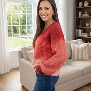 Puede incluir: Un suéter degradado rojo y naranja con cuello en V. El suéter tiene un diseño de punto texturizado y mangas largas. La persona lleva vaqueros azules. El fondo incluye una ventana, un sofá y una estantería.