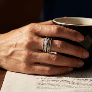 Puede incluir: Un anillo plateado con un diseño multibanda, incrustado con pequeñas piedras brillantes. El anillo se lleva en una mano que sostiene una taza negra. El intrincado diseño del anillo capta la luz. El anillo es una joya.