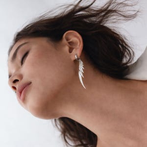 以下が含まれることがあります： 羽のデザインのシルバーイヤリング。イヤリングは、耳に取り付けられた小さなフープに取り付けられています。 羽のデザインは多層で、長く細い形をしています。