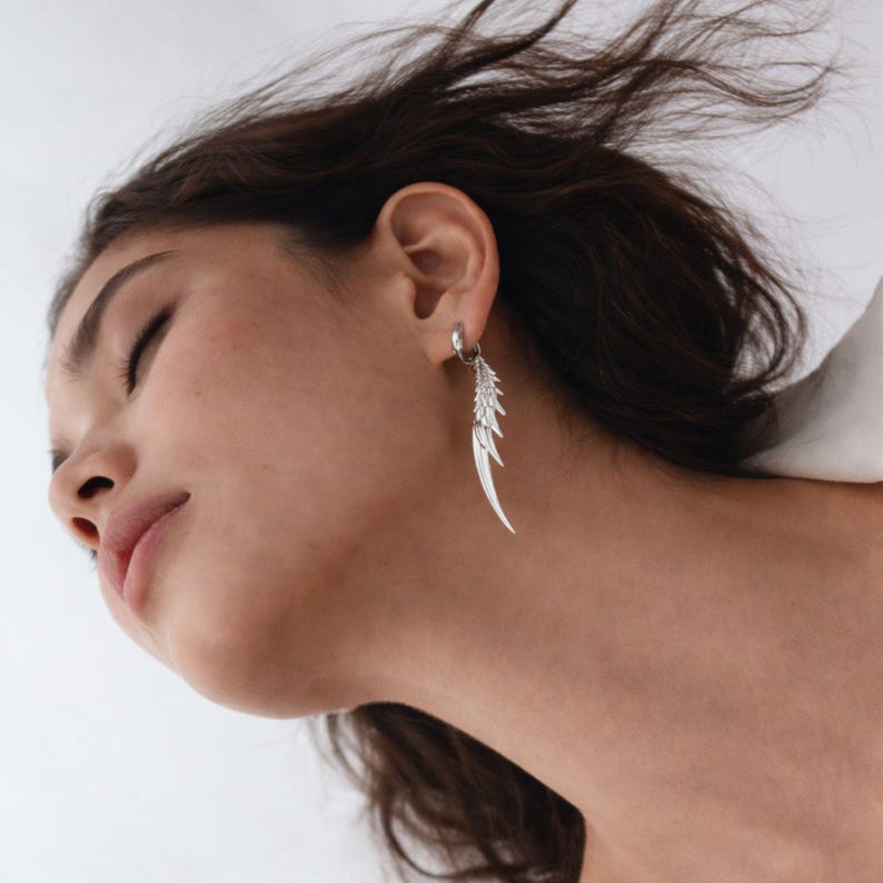 May include: A silver earring with a feather design is shown. The earring is attached to a hoop that is attached to the ear. The feather design has multiple layers and is long and slender.