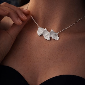 May include: A silver necklace featuring three detailed ginkgo leaf pendants. The necklace is worn around the neck, with the leaves resting on the chest. The leaves have a textured surface and are connected by a delicate chain.