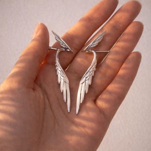 May include: A pair of silver-colored wing-shaped earrings. Each earring features a detailed feather design, with a curved lower section and a smaller wing at the top. The earrings are displayed against a neutral background.