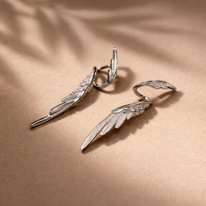 May include: A pair of silver-colored wing-shaped earrings. The earrings have a detailed feather design and a curved post that wraps around the ear. The earrings are displayed on a neutral-toned surface.