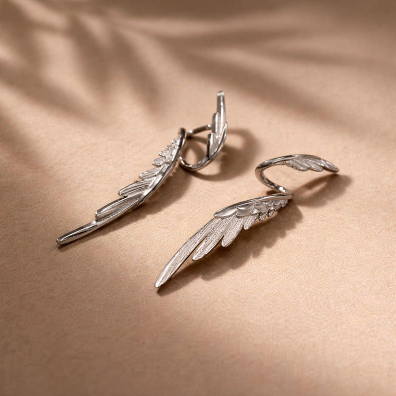 May include: A pair of silver-colored wing-shaped earrings. The earrings have a detailed feather design and a curved post that wraps around the ear. The earrings are displayed on a neutral-toned surface.