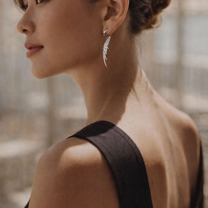 May include: A silver feather-shaped earring is worn by a person. The earring is dangling and has a detailed design. The person is wearing a black top. The background is blurred, with natural light.