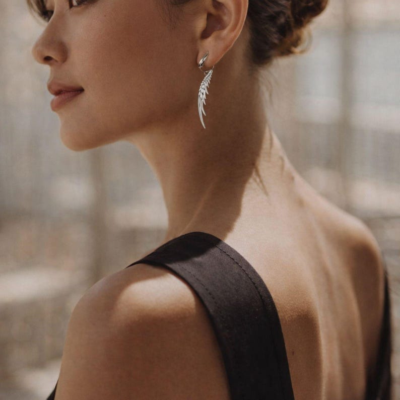 May include: A silver feather-shaped earring is worn by a person. The earring is dangling and has a detailed design. The person is wearing a black top. The background is blurred, with natural light.