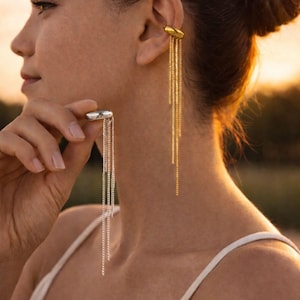 Puede incluir: Una mujer lleva dos pendientes diferentes. Uno es un ear cuff plateado con cadenas colgantes, y el otro es un ear cuff dorado con cadenas colgantes. Los pendientes son un accesorio elegante.