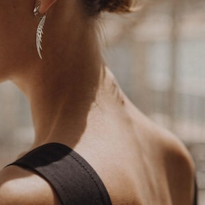 May include: A silver feather earring is shown in a close-up shot. The earring is attached to a small hoop and has a detailed feather design. The background is blurred, with a focus on the earring and the person's neck and shoulder.