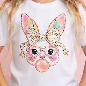 May include: White t-shirt featuring a cartoon bunny face with heart-shaped glasses and a bubblegum bubble. The bunny has a bow with black polka dots and colorful ears. The design is centered on the shirt.