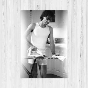 May include: Black and white photograph of a person ironing clothes on an ironing board. The person is wearing a white tank top, shorts, and sunglasses. The ironing board is black and the background is a kitchen.