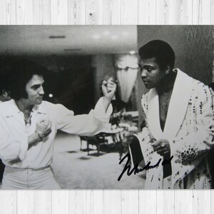 May include: Black and white photograph featuring Elvis Presley and Muhammad Ali. Presley is wearing a white shirt and pants, while Ali is in a white robe. The image is signed by Muhammad Ali.