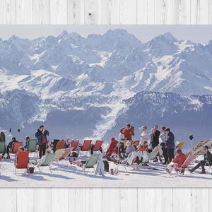 Op de afbeelding: Een vintage foto toont een besneeuwde bergketen met mensen die ontspannen in kleurrijke ligstoelen. De scène omvat een groep mensen in winterkleding, genietend van de zon. De afbeelding roept een gevoel van vrije tijd en de schoonheid van de natuur op.