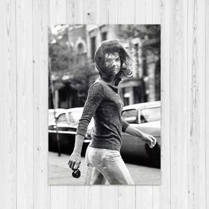 Può includere: Fotografia in bianco e nero di una persona che cammina per una strada cittadina. La persona indossa un top a maniche lunghe e pantaloni, tenendo in mano un piccolo oggetto. Auto ed edifici sono visibili sullo sfondo.