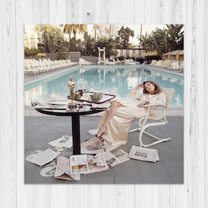 Peut inclure: Photographie vintage d'une femme se relaxant au bord d'une piscine. Elle est assise dans une chaise blanche, vêtue d'une robe crème. Une statuette Oscar est posée sur une table avec des journaux éparpillés. La scène se déroule en extérieur avec des palmiers.