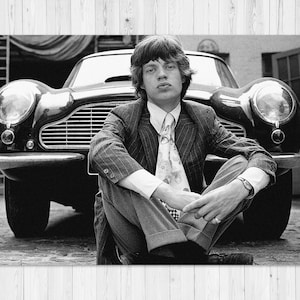 May include: Black and white photograph of a man in a pinstripe suit sitting in front of a classic car. The car has large headlights and a prominent grill. The man is wearing a patterned tie and a wristwatch.