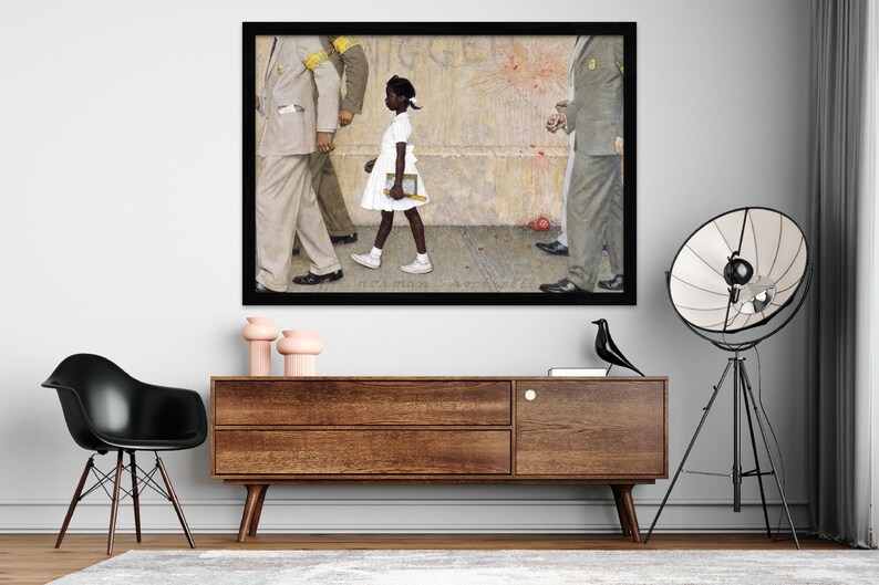 May include: Framed print of a painting depicting a young Black girl in a white dress walking past a group of men in suits. The artwork is set against a textured background with a black frame. The print is displayed above a wooden cabinet.