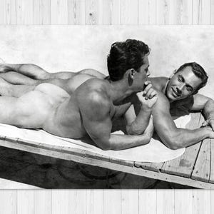 Könnte beinhalten: Schwarz-Weiß-Fotografie von zwei Männern, die auf einer Holzoberfläche liegen. Einer der Männer schaut den anderen an, der lächelt. Das Bild hat einen Vintage-Stil.