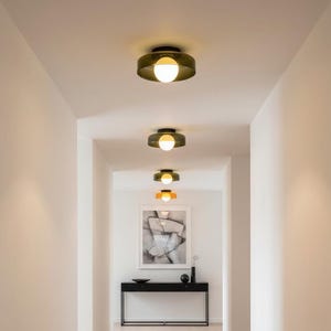 May include: A hallway with recessed lighting fixtures. The fixtures are dark green with a spherical white light. The hallway has white walls, a light wood floor, and a black console table with artwork.
