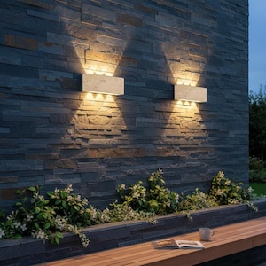 Pode incluir: Duas luminárias de parede retangulares externas iluminam uma parede de pedra cinza escura. As luzes projetam um brilho quente para baixo. Abaixo da parede, há um banco de madeira, com vegetação e um livro visíveis.