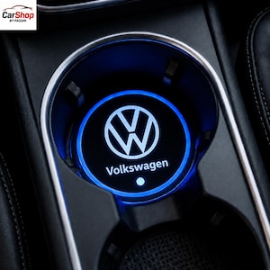 May include: A circular Volkswagen car cup holder coaster featuring a glowing blue LED ring light. The center displays the white Volkswagen logo and the text Volkswagen with a small blue dot below it, set within a black interior console.