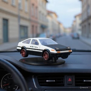 May include: A white and black toy car with red wheels is displayed on a black circular base. The car has a black hood and is positioned on a car dashboard. The background shows a blurred street scene.