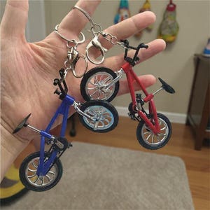 May include: Miniature bicycle keychains in red and blue. Each keychain features a small bicycle with black tires and silver-colored spokes. The handlebars and seat are also black. The keychains are attached to silver-colored key rings.